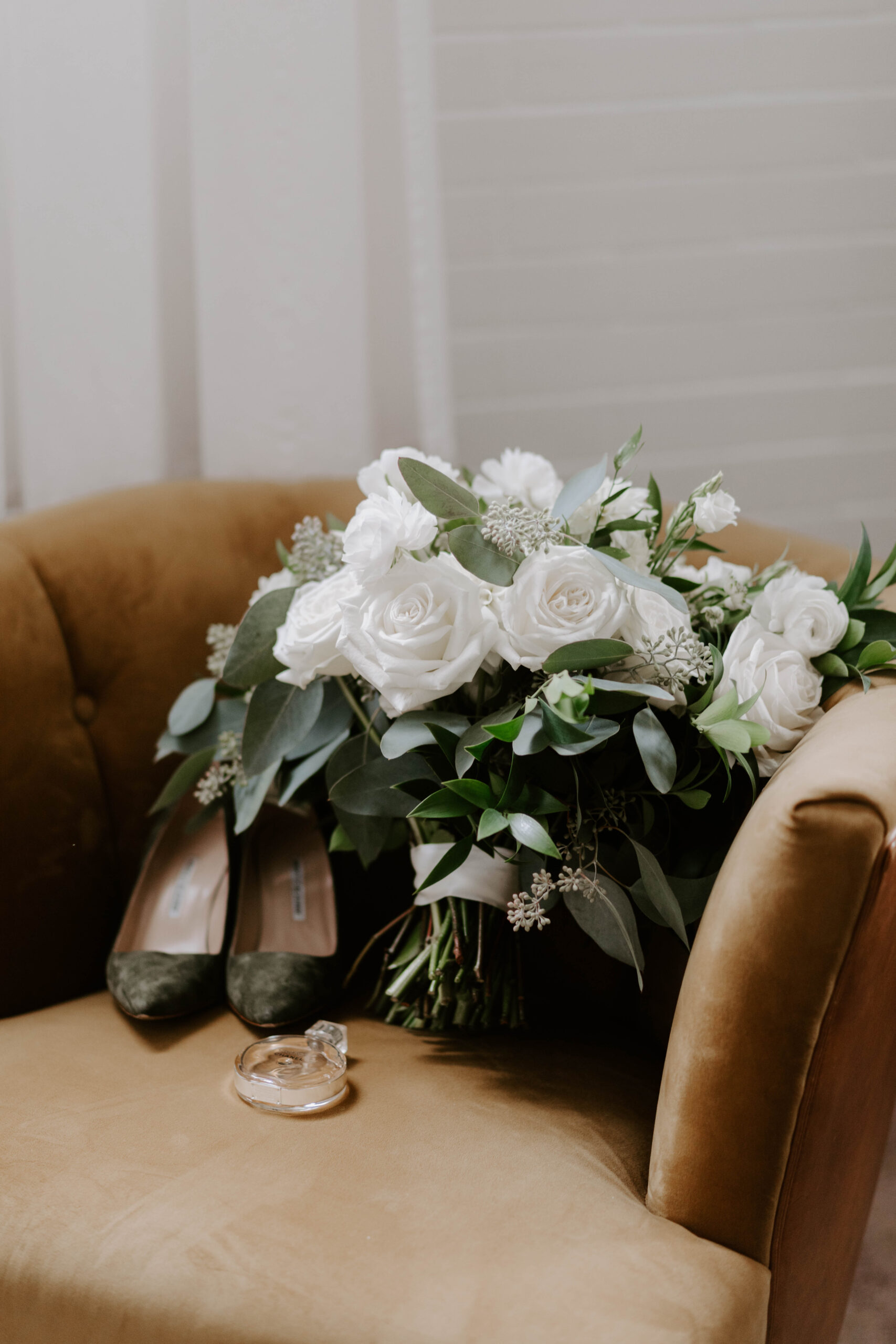 A beautiful wedding bouquet of write roses with greenery, paired with gorgeous mossy green shoes on a velvet chair